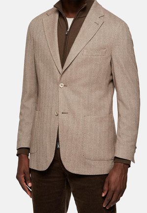 Beige blazer met een visgraatpatroon, met twee knopen, notch revers en zijzakken, gelaagd over een bruine ritsjack aan de binnenkant.