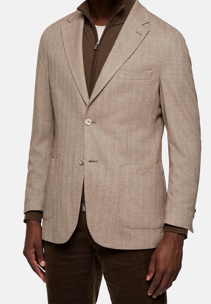 Blazer beige con patrón de espiga, con dos botones, solapas de muesca y bolsillos laterales, superpuesto a una chaqueta interior marrón con cremallera.