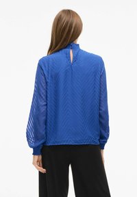 Blusa blu a motivo a zigzag con maniche lunghe trasparenti e collo alto. Presenta un'apertura a keyhole sul retro e polsini elasticizzati.