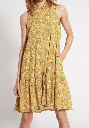 Ærmeløs gul blomsterkjole med høj rynket halsudskæring og løs, flydende pasform, der slutter over knæene, båret af en stående kvinde.