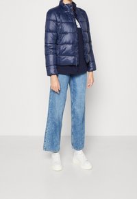 Granatowa kurtka puchowa z wysokim kołnierzem, pikowanym wzorem i zamkiem z przodu, w połączeniu z ciemną górą i jasnoniebieskimi spodniami typu wide-leg.