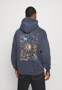 Sudadera azul marino con un gran gráfico en la parte posterior que presenta un sol y diseños circulares. Tejido suave, corte relajado y bolsillo canguro.