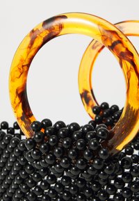 Črne sferične kroglice tvorijo teksturirano površino, ki jo dopolnjujeta dva jantarno marmorirana smolna obroča kot ročaji. Celoten dizajn je kompakten in edinstven.