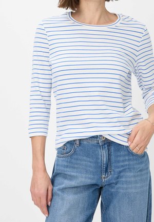 Vrouw draagt een wit shirt met dunne blauwe horizontale strepen en een blauwe spijkerbroek, hand rustend in de zak tegen een witte achtergrond.