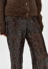 Cardigan tricotat maro, cu închidere cu nasturi, asociat cu pantaloni negri cu paiete, având linii verticale de paiete strălucitoare și un design ajustat.
