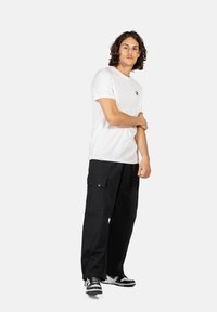 Weißes Baumwoll-T-Shirt mit kleinem Logo, kombiniert mit schwarzen Cargo-Hosen mit seitlichen Taschen und schwarz-weißen Sneakers.