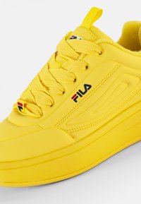 Gul atletik sko med en glatt syntetisk overdel, tykk såle, teksturerte snører, og Fila-logo i svart og rødt på siden.