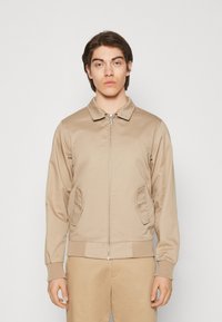 Chaqueta bomber beige con cierre frontal, cuello y bolsillos laterales. Presenta puños y dobladillo acanalados, textura suave y un diseño ligero.