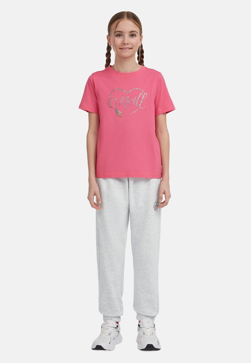 Rožinė medvilninė marškinė su trumpomis rankovėmis, papuošta blizgančia širdies grafika, derinama su pilkais sportiniais šortais ir baltais sportbačiais.