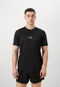 Svart atletisk T-skjorte laget av et lett, pustende stoff, har subtile perforeringer, rund hals og en liten logo på brystet.
