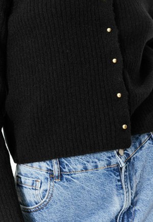 Pull en maille noir avec des boutons dorés sur le côté, associé à un jean en denim bleu clair avec une poche avant et des détails de couture.