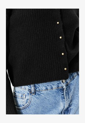 Pull en maille noir avec des boutons dorés sur le côté, associé à un jean en denim bleu clair avec une poche avant et des détails de couture.