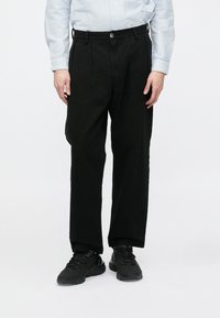 Zwarte broek met een ontspannen pasvorm, voorzien van een gladde textuur, zijzakken en een knoopsluiting. Draag ze met zwarte sneakers.