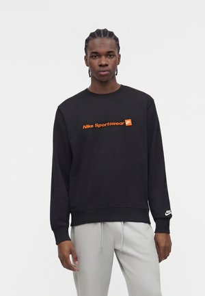 Jeune homme portant un sweat-shirt noir Nike Sportswear avec un logo orange et un pantalon gris clair, debout devant un fond blanc uni.