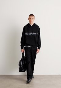 Svart hettegenser med "HUGO" logo, kombinert med svarte cargo-bukser. Holder en svart skinnveske, står modellen foran en nøytral bakgrunn.