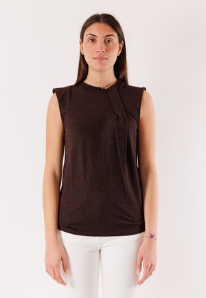 Donna con lunghi capelli castani che indossa un top marrone scuro senza maniche con dettaglio drappeggiato e pantaloni bianchi, in piedi davanti a uno sfondo neutro.