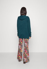 Teal huva kardigan i strukturerat material, parat med utsvängda byxor med blommönster som har röda, lila och gula blommor på en grå bakgrund.