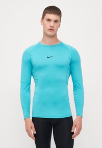 Langærmet atletisk skjorte i lys turkis stof med en glat tekstur, der har et sort Nike-logo på midten af brystet.