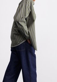 Zielona koszula w paski z białymi i fioletowymi liniami, o luźnym kroju z guzikami, noszona z szerokimi, ciemnoniebieskimi spodniami.