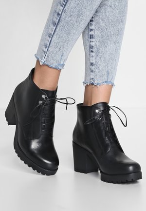 Black leather ankle boots with block heels and laces worn with light blue frayed hem jeans on a white background.