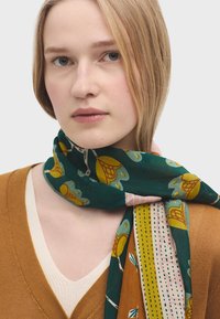 Grønne mønstrede tørklæde med blomsterdesigns og striber, lavet af letvægtsstof. Båret over en brun sweater og en cremefarvet top.