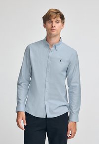 Ljusblå skjorta med krage och långa ärmar. Har en liten svart logotyp på bröstet. Slät tygtextur.