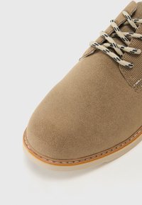 Zapato de ante color beige con puntera redondeada, costuras en contraste y cordones texturizados beige sobre un panel de malla oscura. Suela de goma con detalles a rayas.