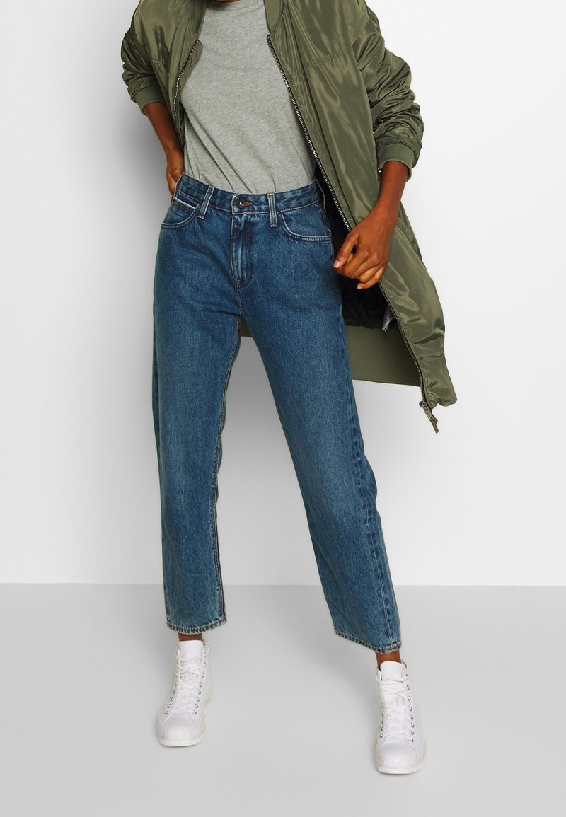 Personne portant une veste bomber vert olive, un t-shirt gris, un jean taille haute bleu et des baskets montantes blanches, debout devant un fond blanc.