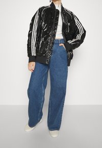 Černá, lesklá bomber bunda s bílými pruhy, žebrovanými manžetami a límcem. Skombinována s vysokými modrými džíny s širokými nohavicemi a světlými teniskami.