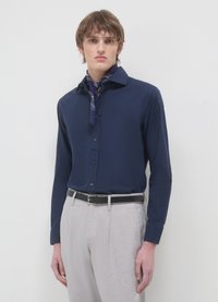 Marineblaue Hemd mit Knöpfen und spitzem Kragen, kombiniert mit hellgrauen, strukturierten Hosen und einem dunkelblauen, gemusterten Schal um den Hals.