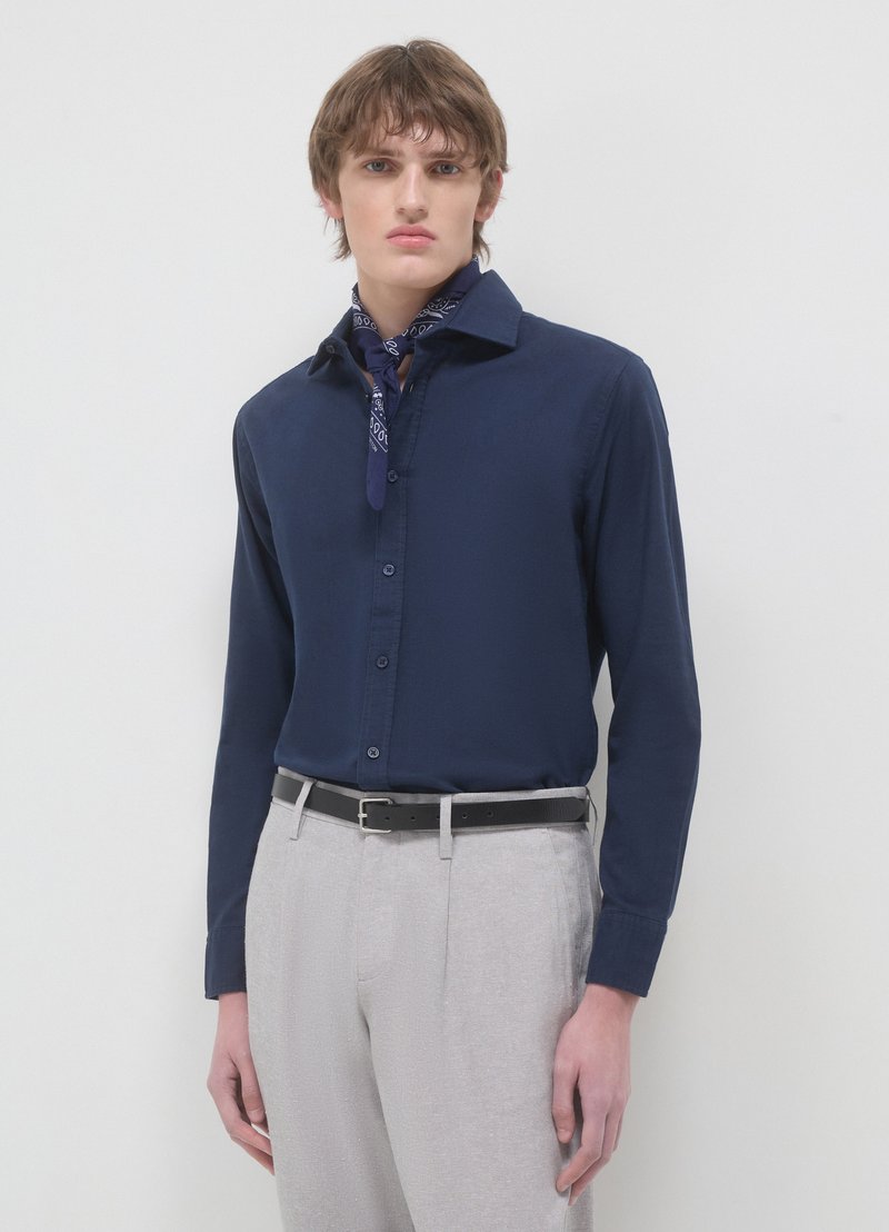 Marineblaue Hemd mit Knöpfen und spitzem Kragen, kombiniert mit hellgrauen, strukturierten Hosen und einem dunkelblauen, gemusterten Schal um den Hals.