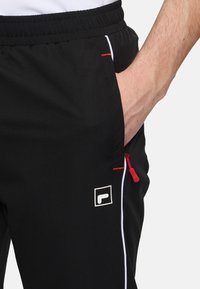 Czarne spodnie sportowe z czerwonym zamkiem błyskawicznym, białymi obszyciami i logo na udzie. Wykonane z gładkiego, lekkiego materiału.