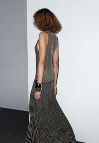 Una modella con capelli ricci indossa un vestito a righe nere con un taglio laterale, in piedi contro uno sfondo minimalista.