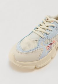 Baskets Lacoste en crème et bleu clair. Caractéristiques : panneaux en maille, accents texturés et détail du logo en rouge. Semelle en caoutchouc, design à lacets.