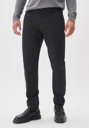 Pantalones negros de tejido entrelazado, ajustados, con trabillas para cinturón, bolsillos frontales y una textura suave. Llevados con zapatos negros tipo slip-on.