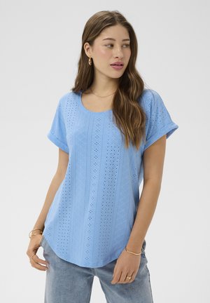 Vrouw met lang bruin haar kijkt naar links en draagt een lichtblauwe top met opengewerkte korte mouwen en lichtblauwe jeans, gecombineerd met gouden sieraden.