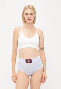 Bela braletka z nastavljivimi naramnicami in gladko teksturo, v kombinaciji z svetlo vijoličastimi hlačkami visokega pasu, ki imajo bordo rdeč logotip Calvin Klein.