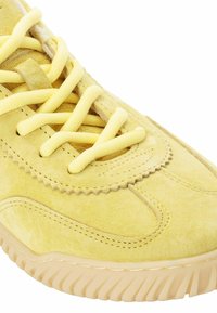 Zapatilla de ante amarilla con cordones a juego, parte superior texturizada, detalles de costura en zigzag y una suela gruesa de goma con estrías.
