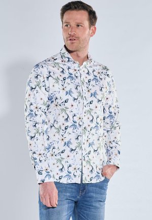 Man wearing a long-sleeve white shirt with blue and green floral patterns, paired with blue jeans, standing against a plain light background.