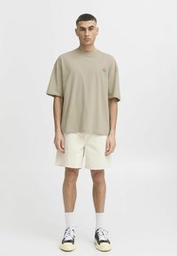 T-shirt oversize kaki avec un design de soleil imprimé, associé à un short beige clair. Porté avec des baskets noires et blanches et des chaussettes blanches.