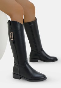 Botas de cuero negro hasta la rodilla con cremallera lateral, tacón plano y hebilla decorativa en tono dorado en el lado lateral. Acabado suave, diseño elegante.