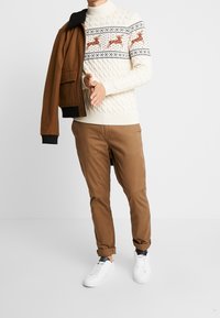 Stickad krämfärgad tröja med kabelmönster och renar, brun jacka, beige byxor och vita sneakers, händerna poserade framåt.