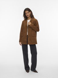 Mujer con blazer marrón con botones, top blanco, jeans negros de pierna ancha y zapatos marrones de punta, posando contra un fondo blanco.