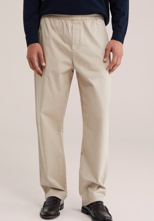 Homme portant un pantalon beige à taille élastique et jambes droites, et des mocassins marron foncé, debout les mains relâchées le long du corps.