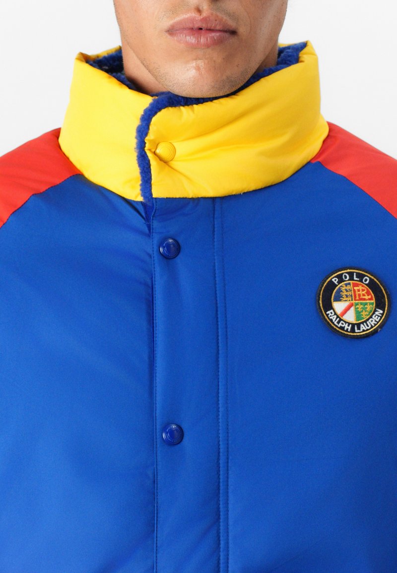 Niebieska puchowa kurtka z żółtym kołnierzem i czerwonymi ramionami. Posiada niebieskie zapięcie na guziki i okrągłą łatkę z logo na piersi. Miękka tekstura.