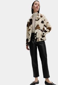 Umělé kožešinové sako s hnědým a bílým vzorem kravské kůže, zapínání na zip a boční kapsy, kombinované s černými těsnými kalhotami a šněrovacími botami.