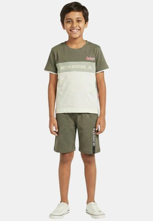 Garçon souriant portant un t-shirt "British Heritage UK" vert et beige, un short vert avec logo sur le côté, et des baskets blanches, debout devant un fond uni.