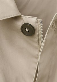 Lähikuva beigenvärisestä kankaasta, jossa on tumma nappi ja osittain näkyvä hopeinen vetoketju vaatteen kauluksessa.