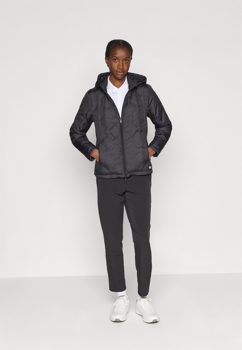 Giacca a vento nera con cappuccio, caratterizzata da un motivo trapuntato, chiusura con zip e tasche laterali, abbinata a pantaloni scuri e sneakers bianche.