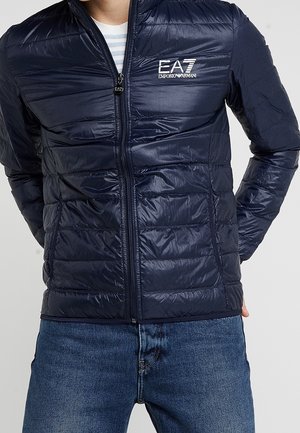 Mand iført navyblå quiltet EA7 Emporio Armani jakke med lynlås og blå jeans, hænderne bag ryggen, foran neutral baggrund.